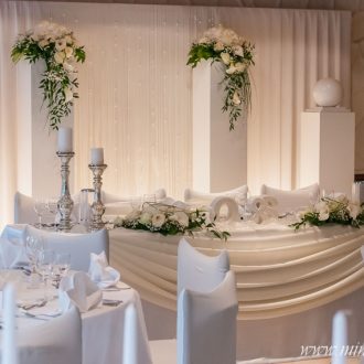 Brauttisch mit Hintergrund. Weisse Säulen mit 2 Blumengestecken und 2 weißen Kugeln, 2 Brauttischgestecken und 4 bauernsilberne Kerzenständer